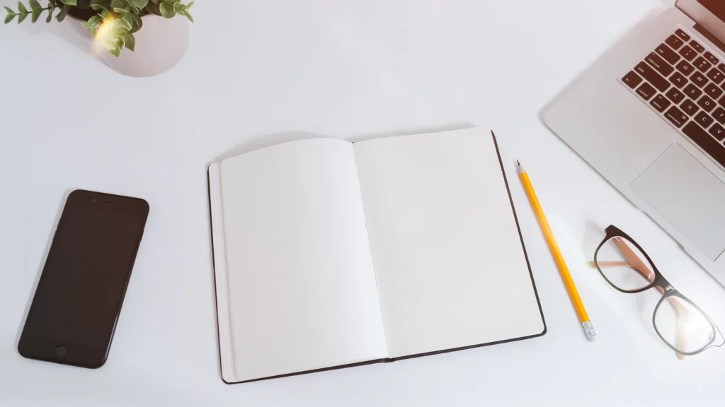 Bureau blanc vu du dessus avec un téléphone intelligent, un carnet ouvert, un crayon, des lunettes et un ordinateur portable