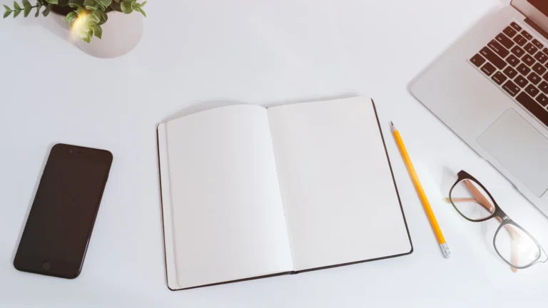 Bureau blanc vu du dessus avec un téléphone intelligent, un carnet ouvert, un crayon, des lunettes et un ordinateur portable