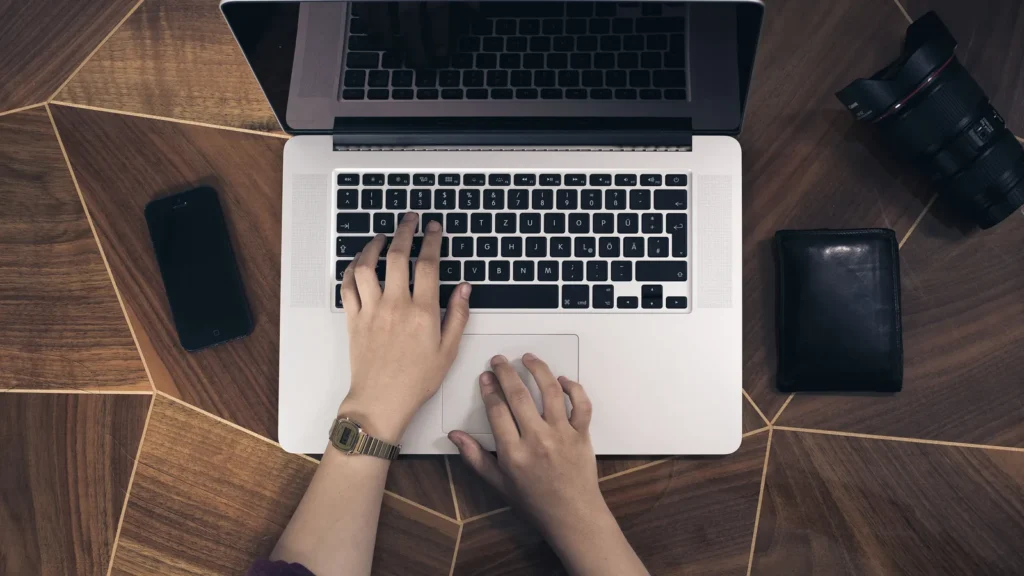 Mains posées sur le clavier d'un ordinateur portable, vues du dessus, sur une table en bois avec un téléphone et un portefeuille