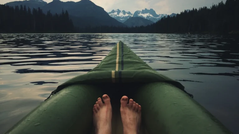 Pieds nus d'une personne dans un canot vert sur un lac calme, devant des montagnes et une forêt de conifères
