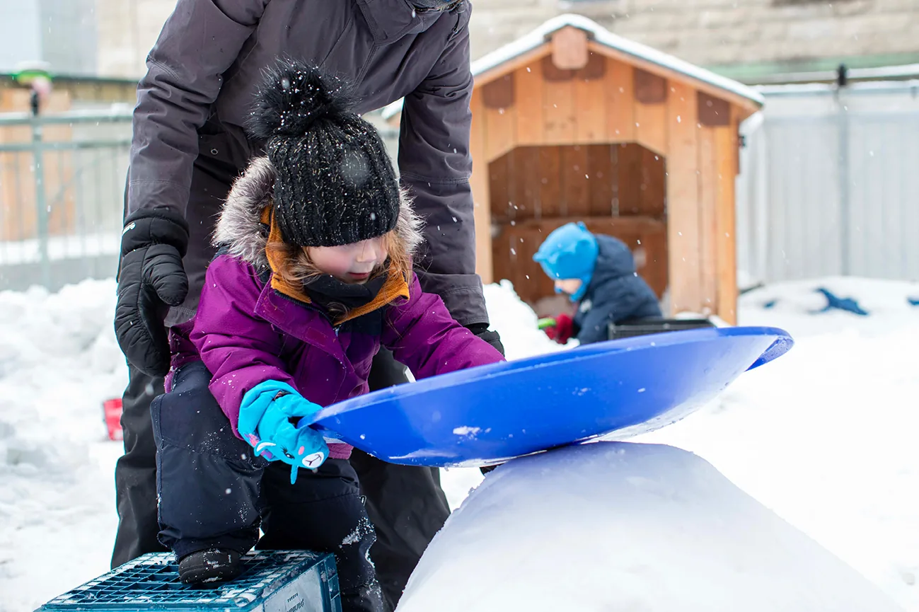 Enfant en habit de neige qui joue avec une pelle bleue près d'une éducatrice
